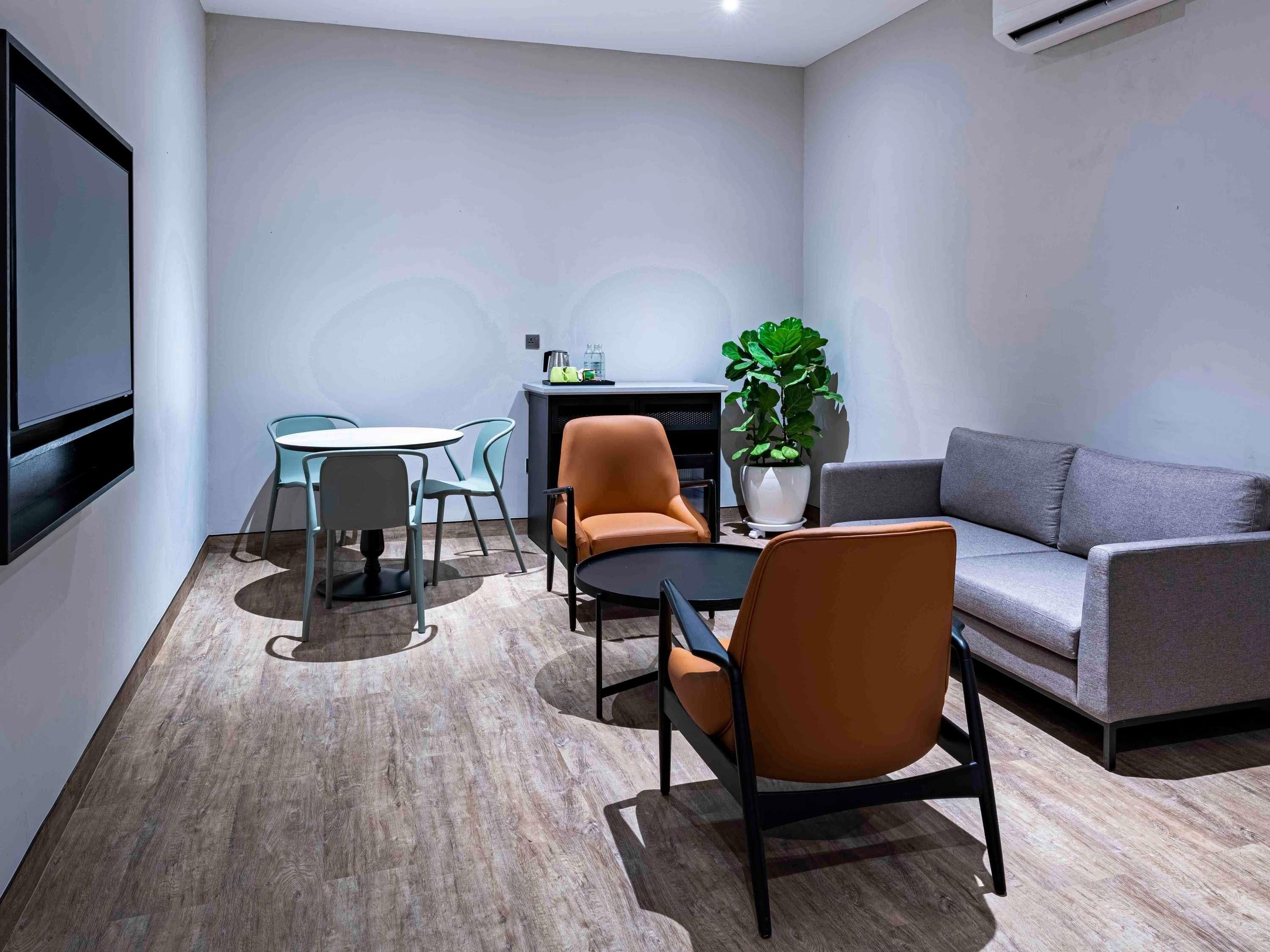 A modern room with a grey sofa, a round table, and two chairs. There's a potted plant and a small table with a kettle and cups.