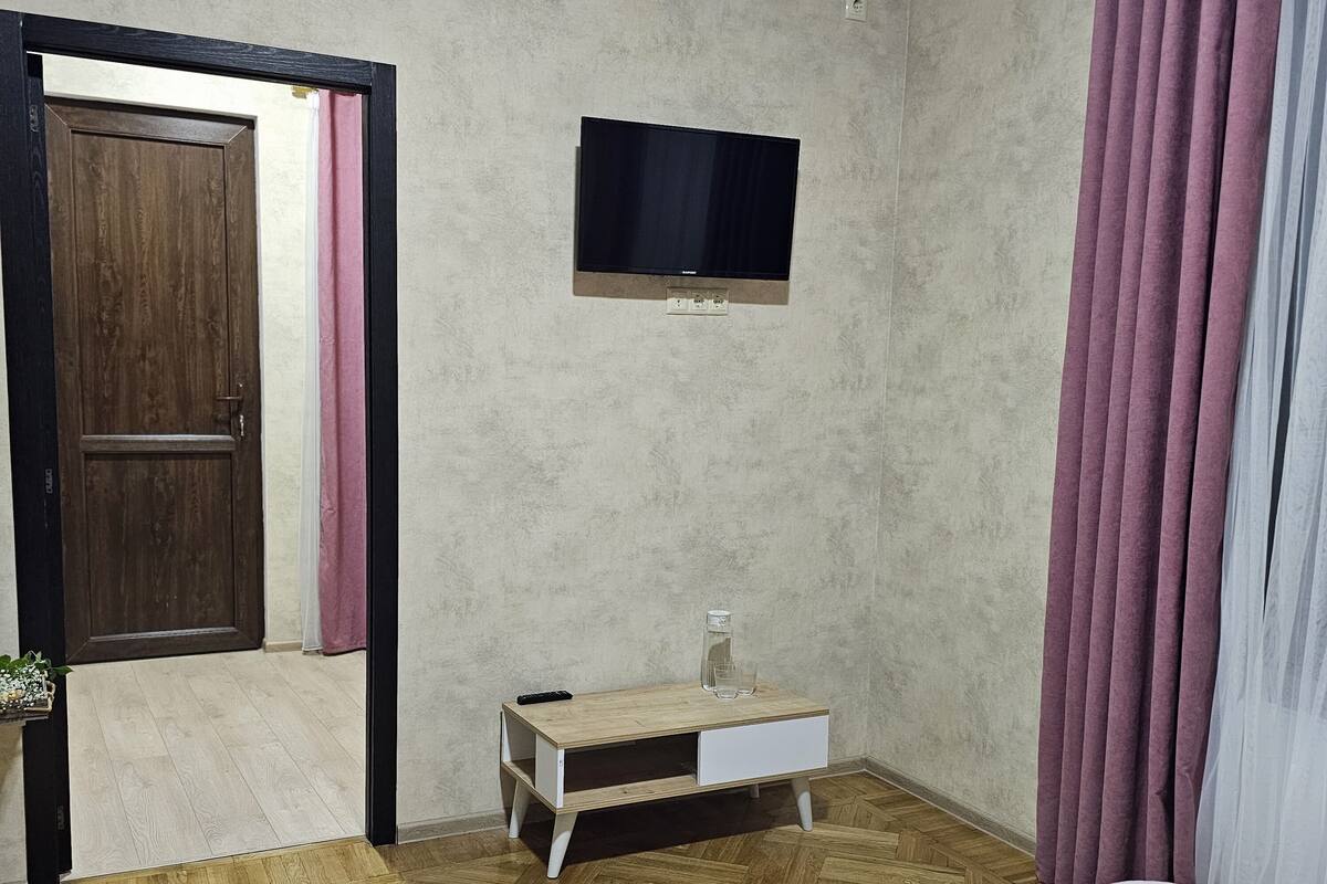 Quarto com piso de madeira, uma cama, uma pequena mesa de cabeceira de madeira com um copo, uma televisão na parede e uma porta com cortinas cor-de-rosa.