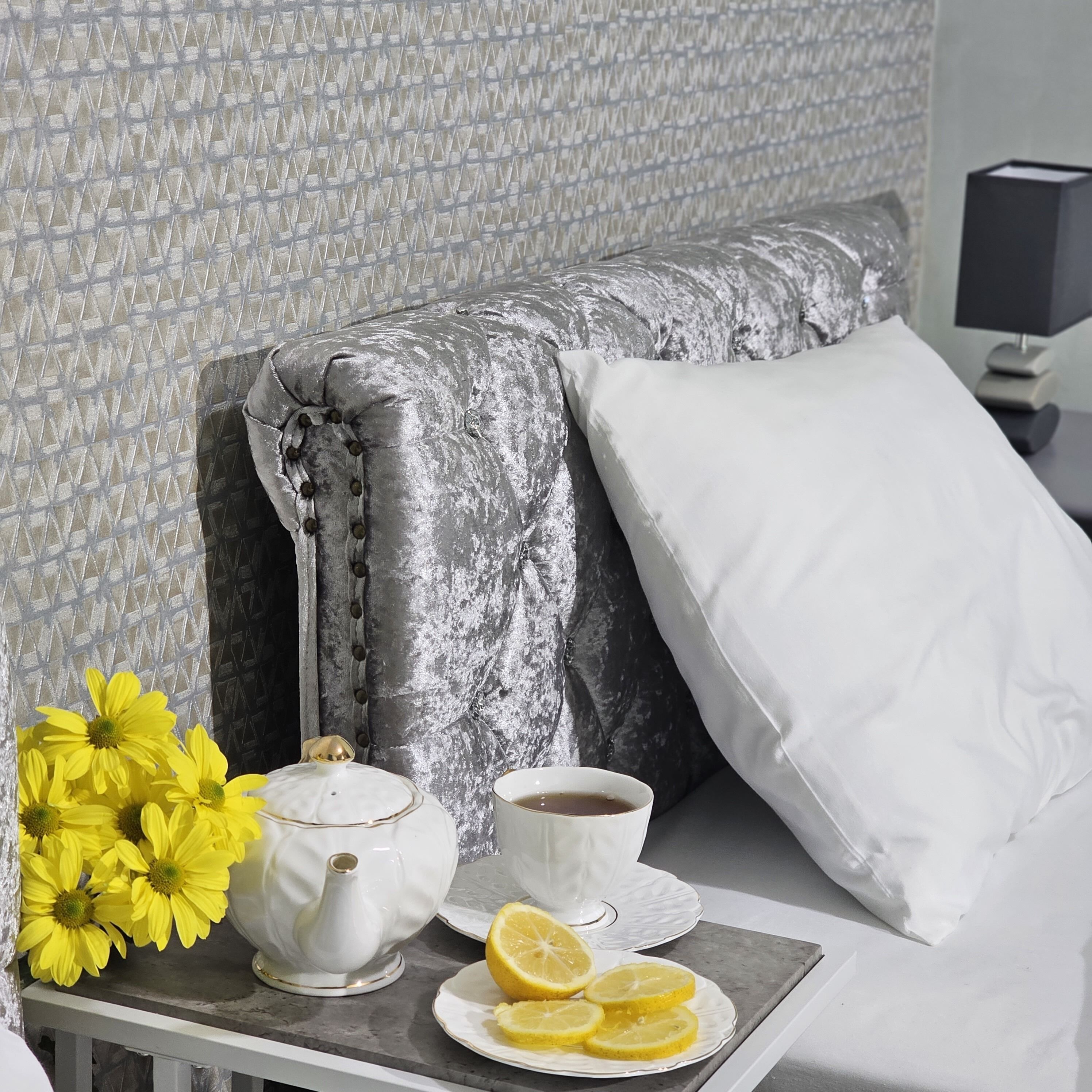 A neatly made bed with a grey upholstered headboard, a white pillow, and a tray table with a teapot, cup, and lemon slices.