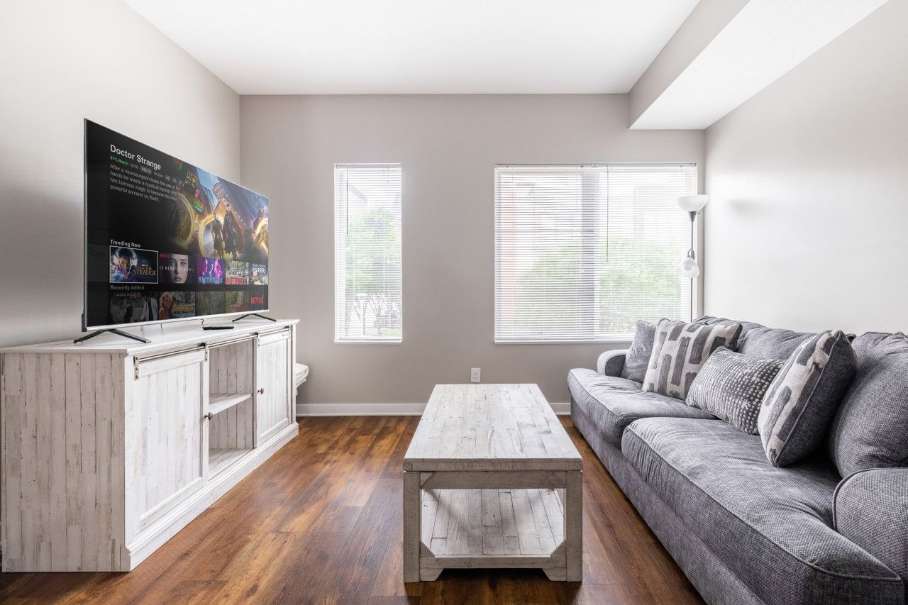 A living room with a TV on a white cabinet, a gray sofa, and two windows with blinds.