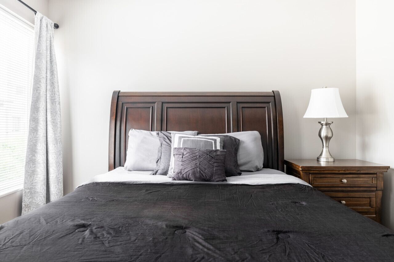 A neatly made bed with a dark headboard, a bedside lamp, and a window with curtains.