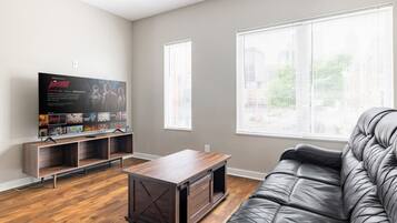A living room with a flat-screen TV, wooden entertainment unit, a leather sofa, and large windows with blinds.
