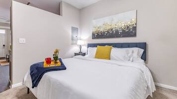 A hotel room with a bed, a nightstand, a lamp, a tray with a red cup and a plant, and a painting on the wall.