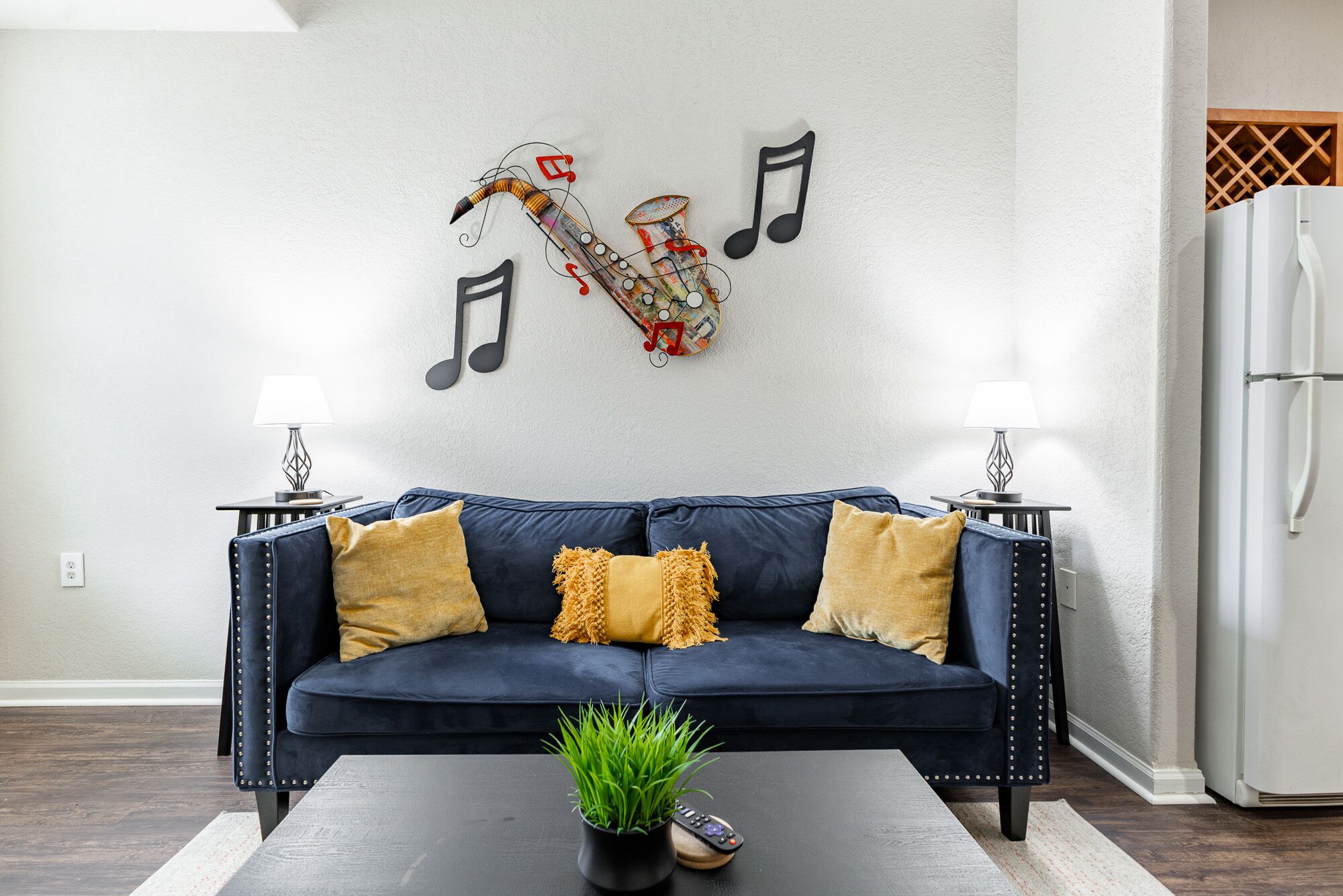 A living room with a dark blue sofa, two yellow cushions, a small potted plant, and a wall-mounted guitar decoration.