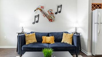 A living room with a dark blue sofa, two yellow cushions, a small potted plant, and a wall-mounted guitar decoration.