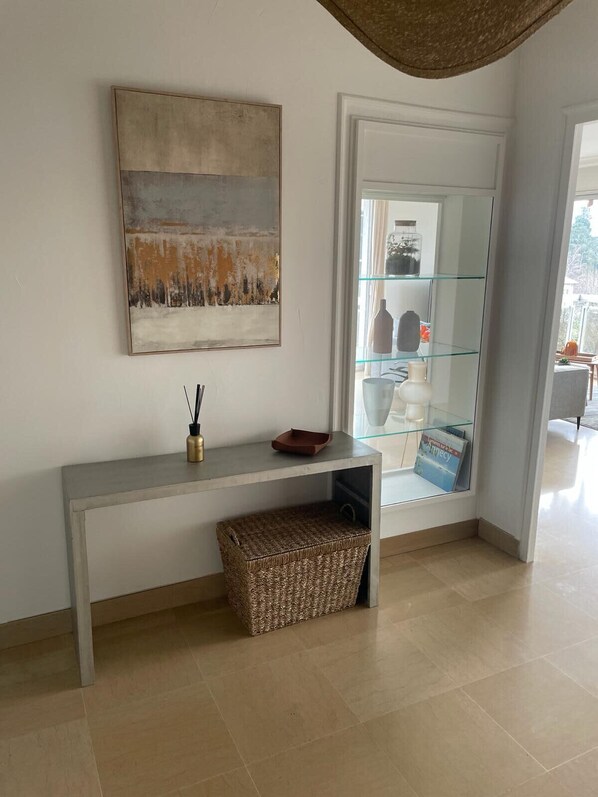 Un soggiorno moderno con un tavolo consolle grigio, un cesto intrecciato, un quadro astratto incorniciato e una vetrina in vetro con oggetti decorativi.