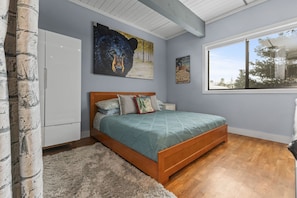A bedroom with a wooden bed, a fluffy rug, a painting of a bear, a window with a view of trees, and a white wardrobe.