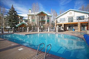 A swimming pool with a wooden deck, surrounded by buildings with balconies and a snow-covered tree.