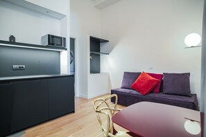 A modern kitchen with a microwave, a built-in oven, and a small refrigerator. There is a dining table with a red tablecloth and a single chair. A dark-colored sofa with red and purple cushions is against the wall. - Youroom Margutta (Rome)