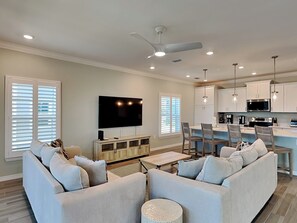 A modern living room with a flat-screen TV, light-colored sofas, and a dining area with a kitchen in the background.
