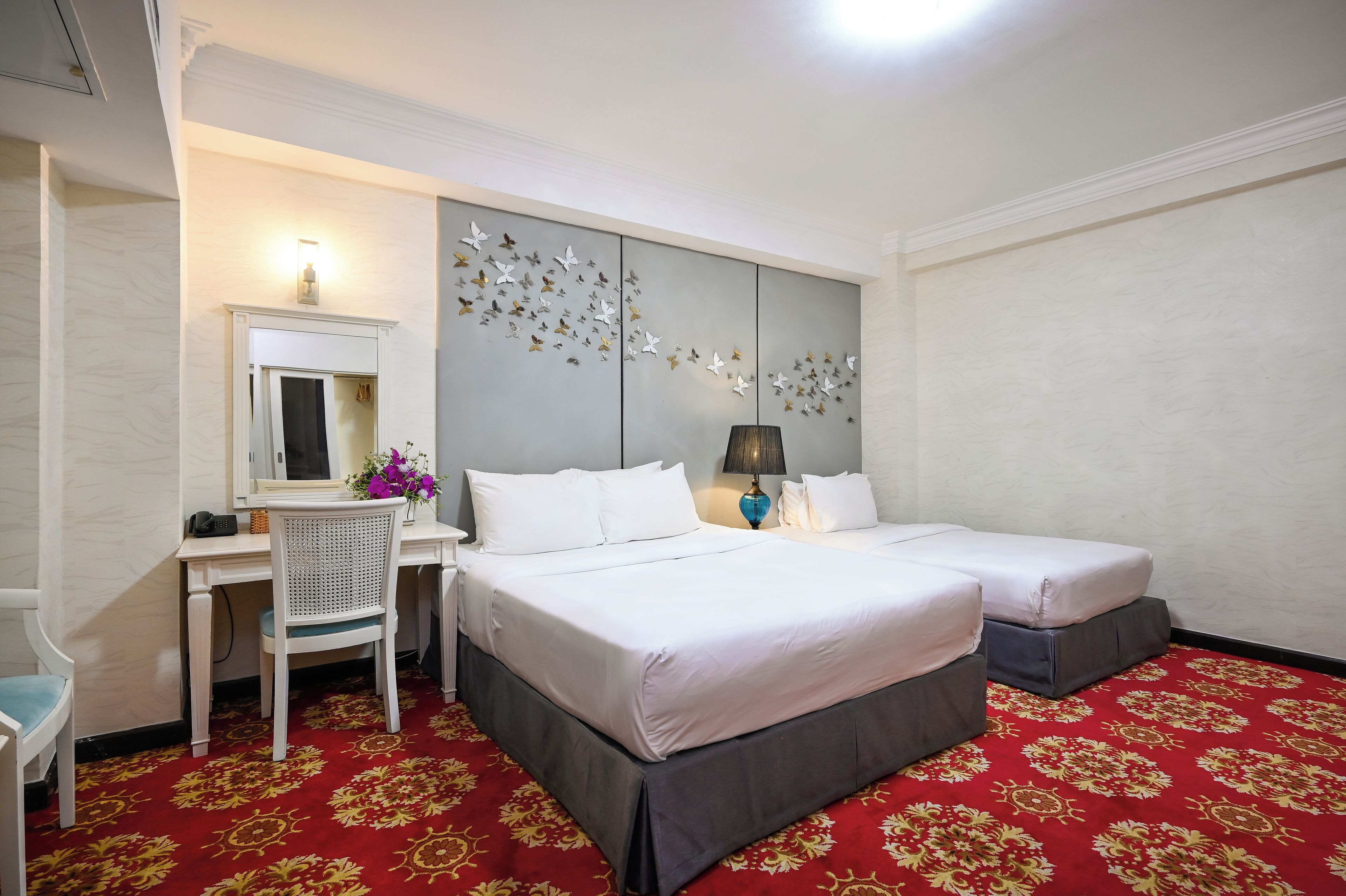 A hotel room with a bed, two bedside tables, a desk with a chair, a lamp, a mirror, and a decorative wall panel.