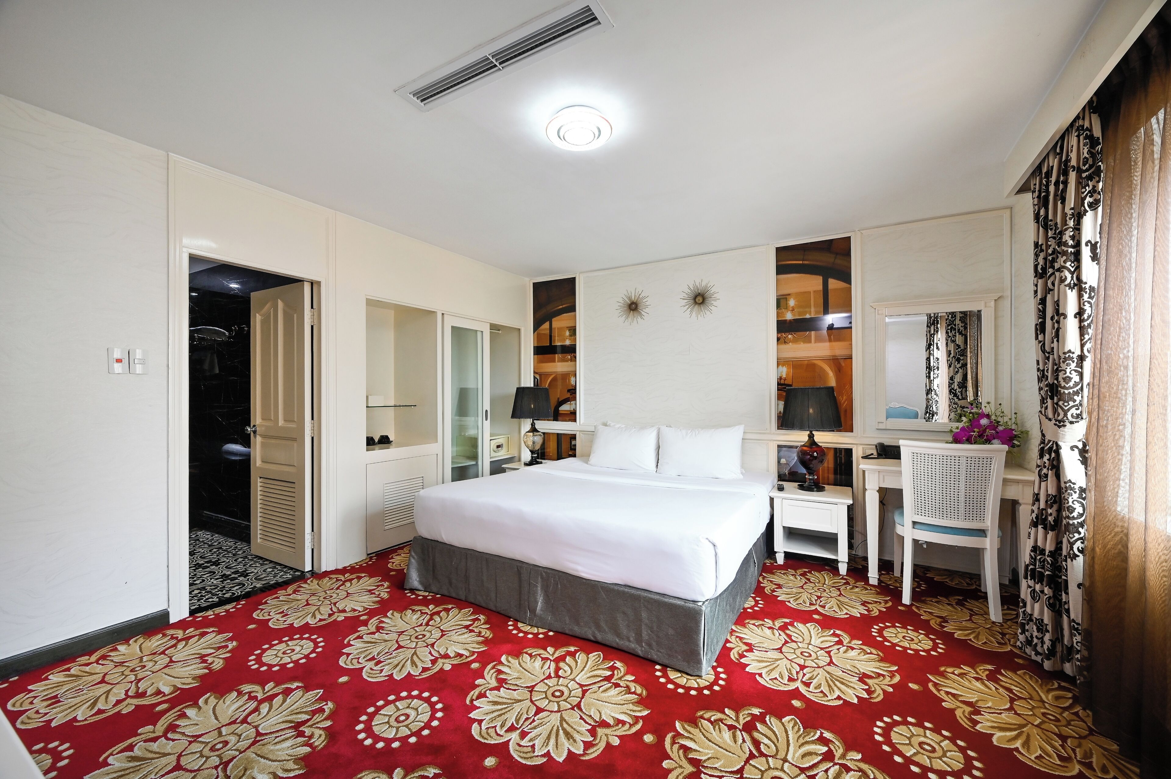 A hotel room with a large bed, a desk, a chair, a mirror, and a patterned carpet.