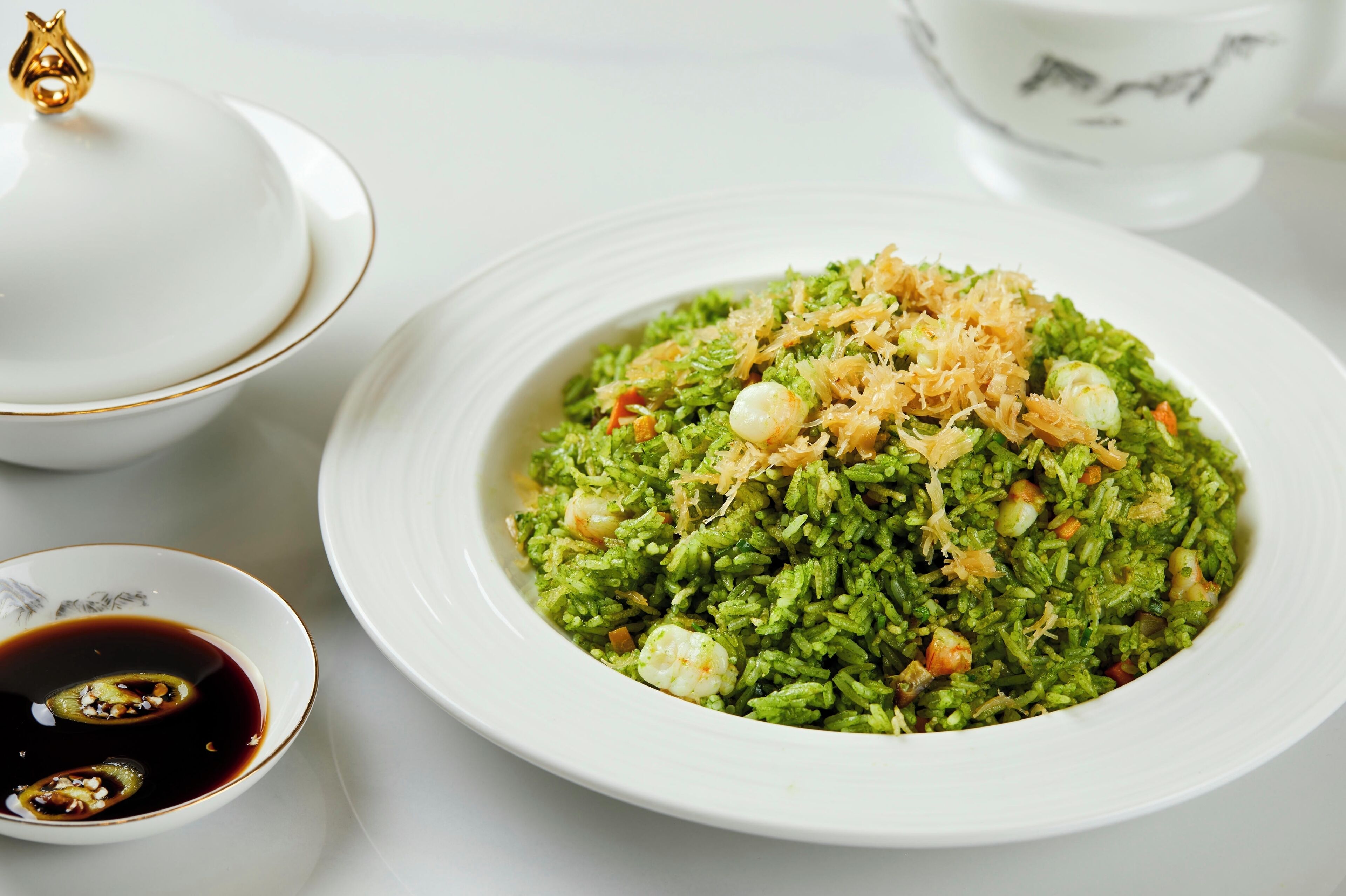 A bowl of green rice with shrimp and fried onions, accompanied by a small bowl of sauce.
