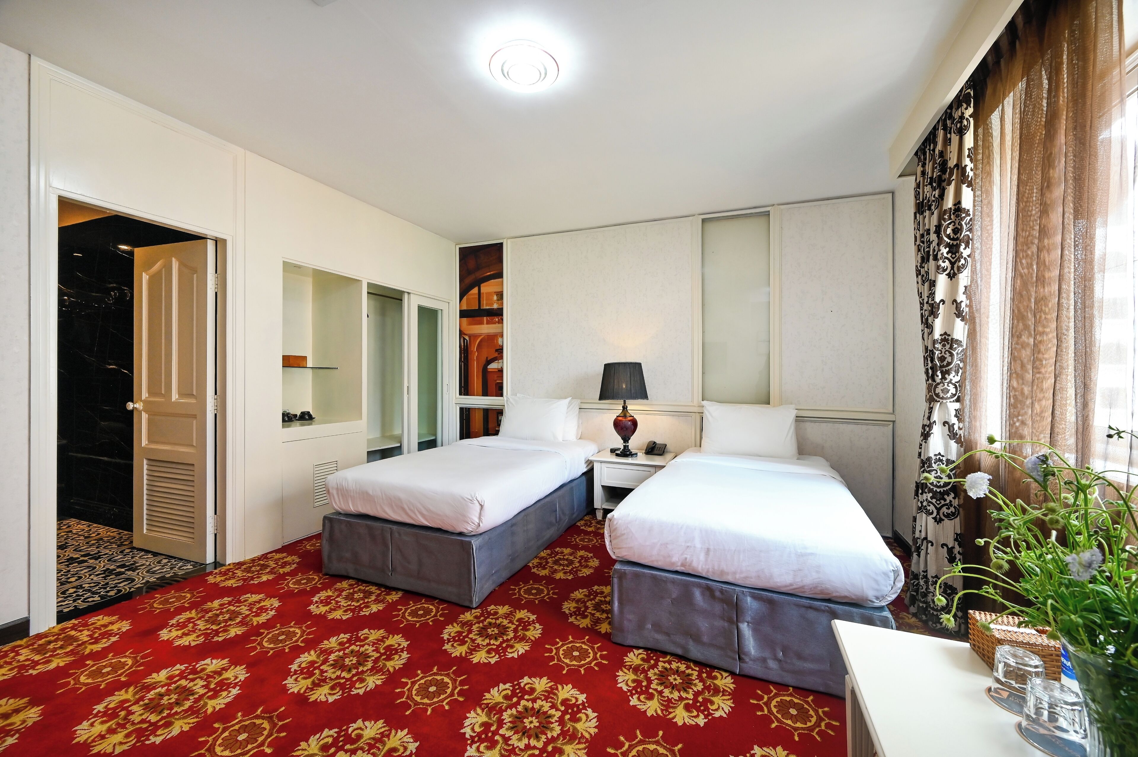 Una habitación de hotel con dos camas, un escritorio, una lámpara y un ventanal con cortinas.