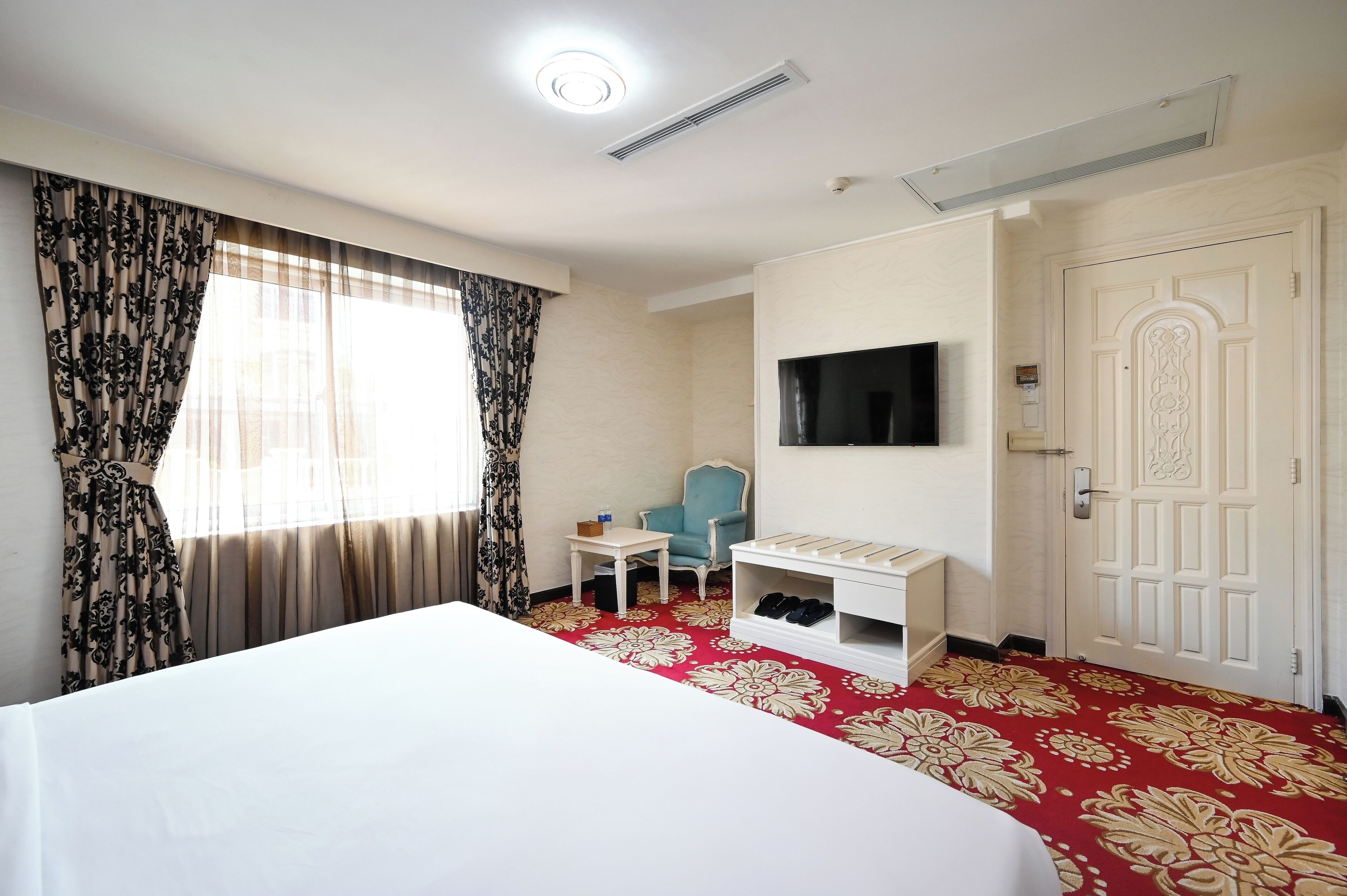 A hotel room with a large bed, a television, a small table, a chair, and a shoe rack.