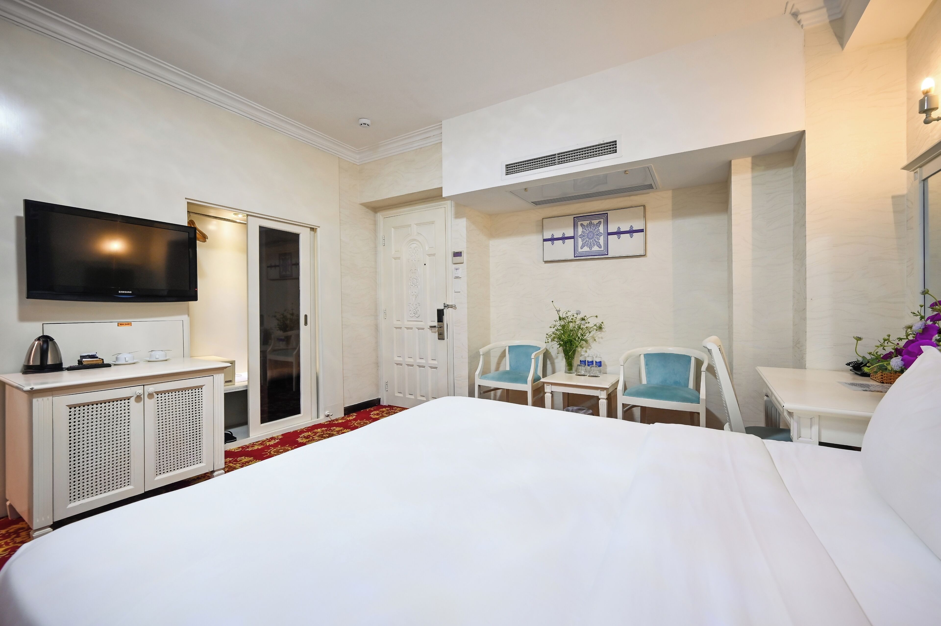 A hotel room with a large bed, a television, a desk with a chair, a small table with a chair, and a door.