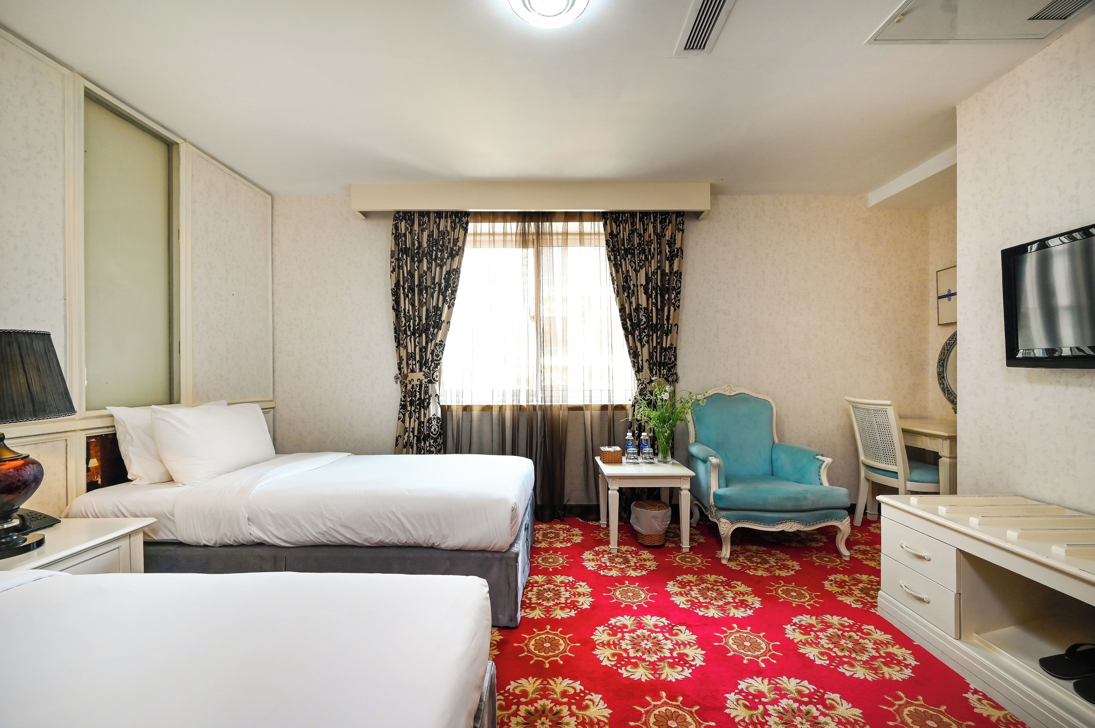 Ein Hotelzimmer mit zwei Betten, einem roten Teppich, einem kleinen Tisch mit Pflanze, einem blauen Sessel und einem Fernseher an der Wand.