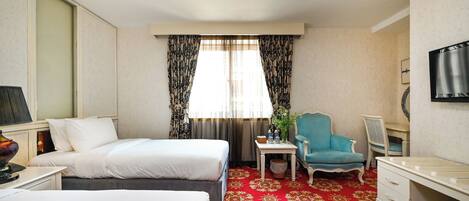 Ein Hotelzimmer mit zwei Betten, einem roten Teppich, einem kleinen Tisch mit Pflanze, einem blauen Sessel und einem Fernseher an der Wand.