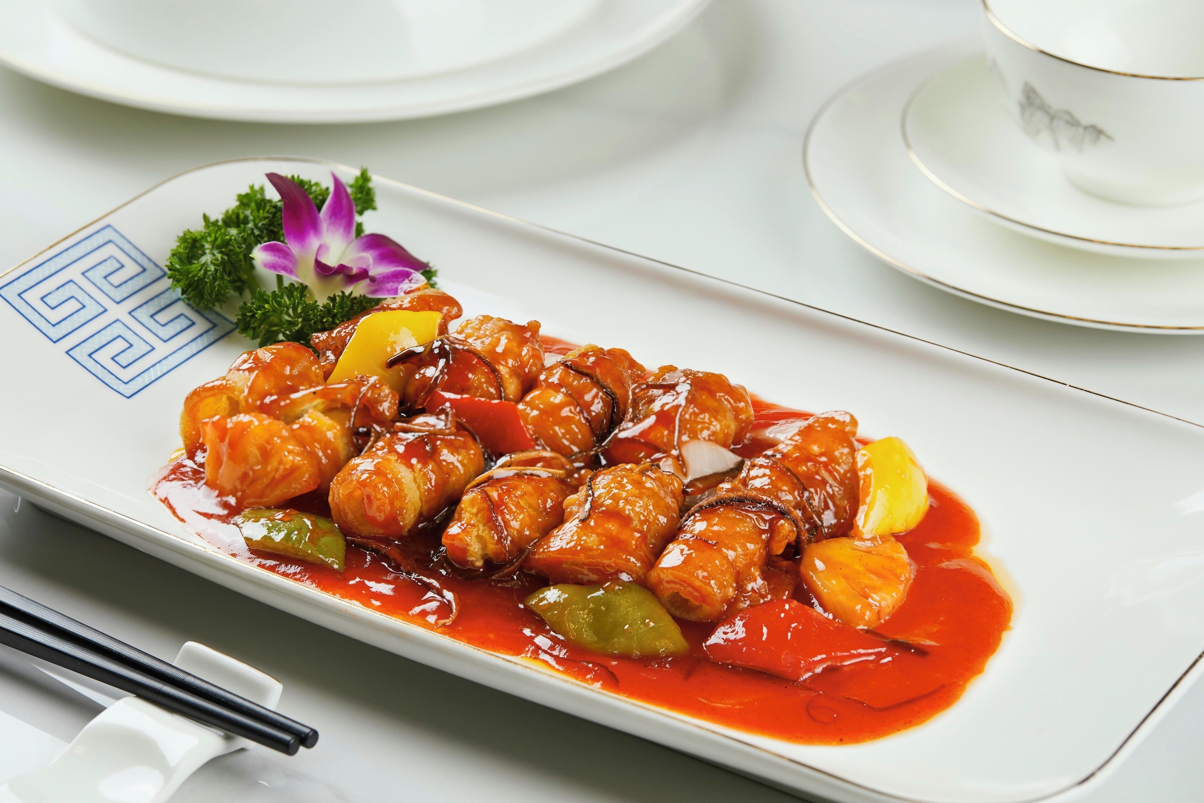 A rectangular plate with a dish of glazed meat and vegetables, accompanied by a small purple flower and green garnish.