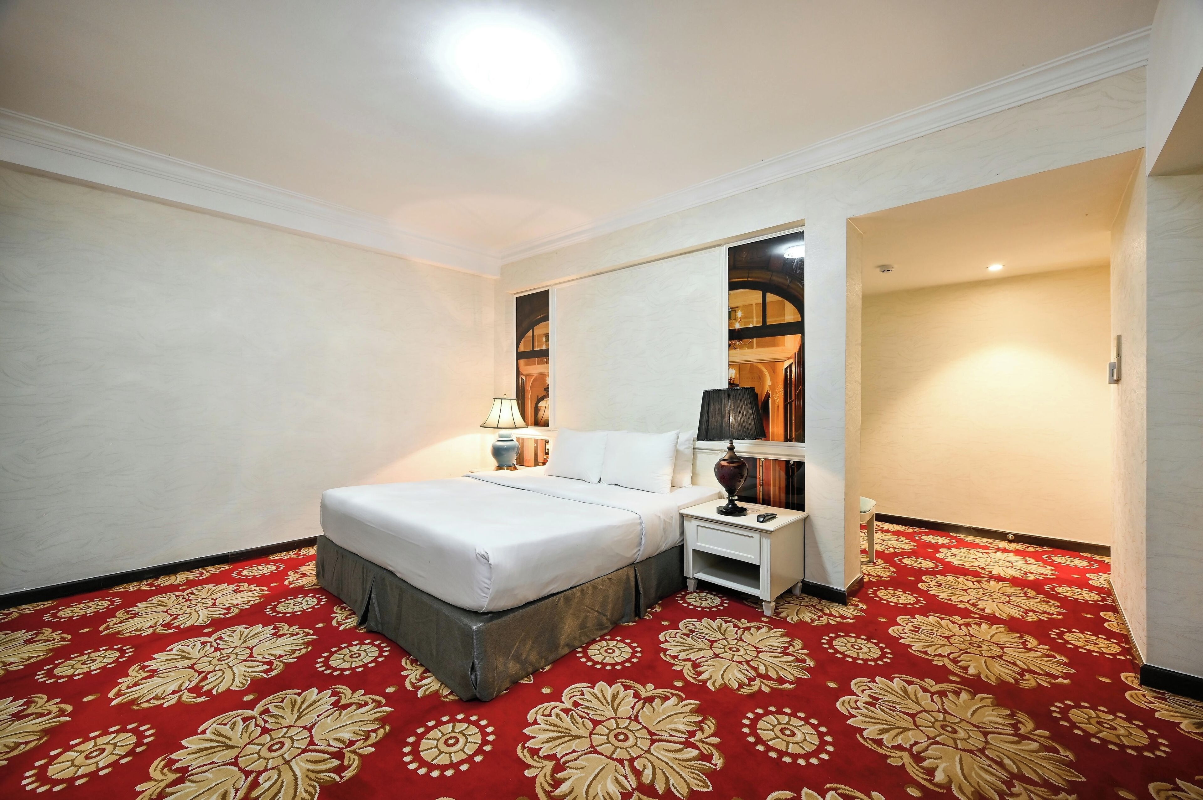 A hotel room with a large bed, a nightstand, a lamp, and a red carpet with a floral pattern.