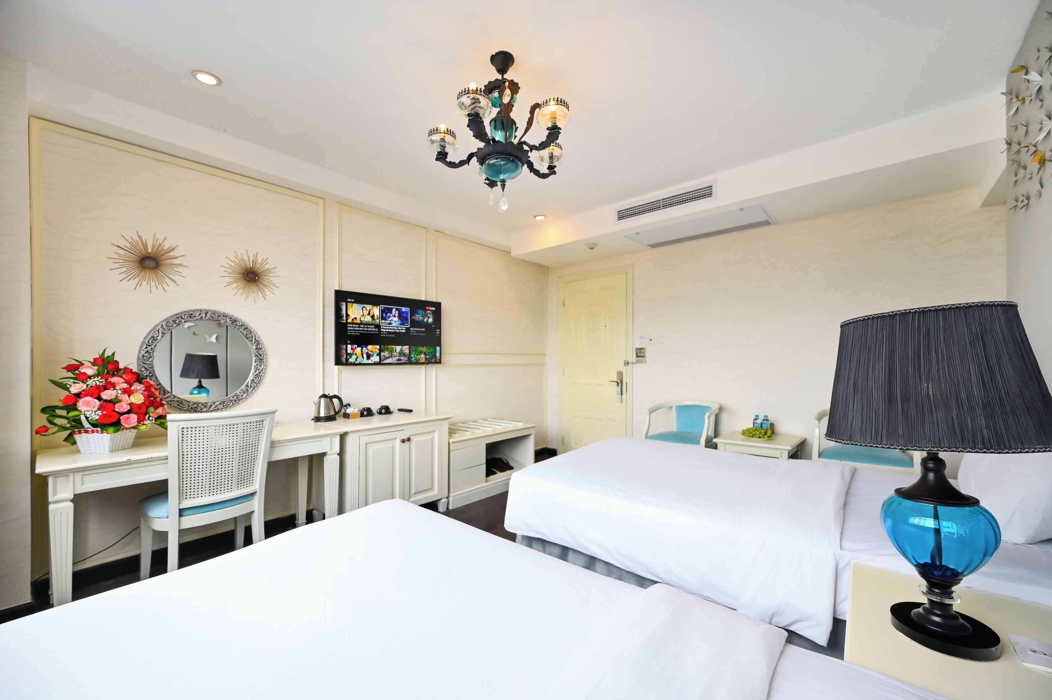 A hotel room with a desk, two beds, a chandelier, a TV, and a lamp.