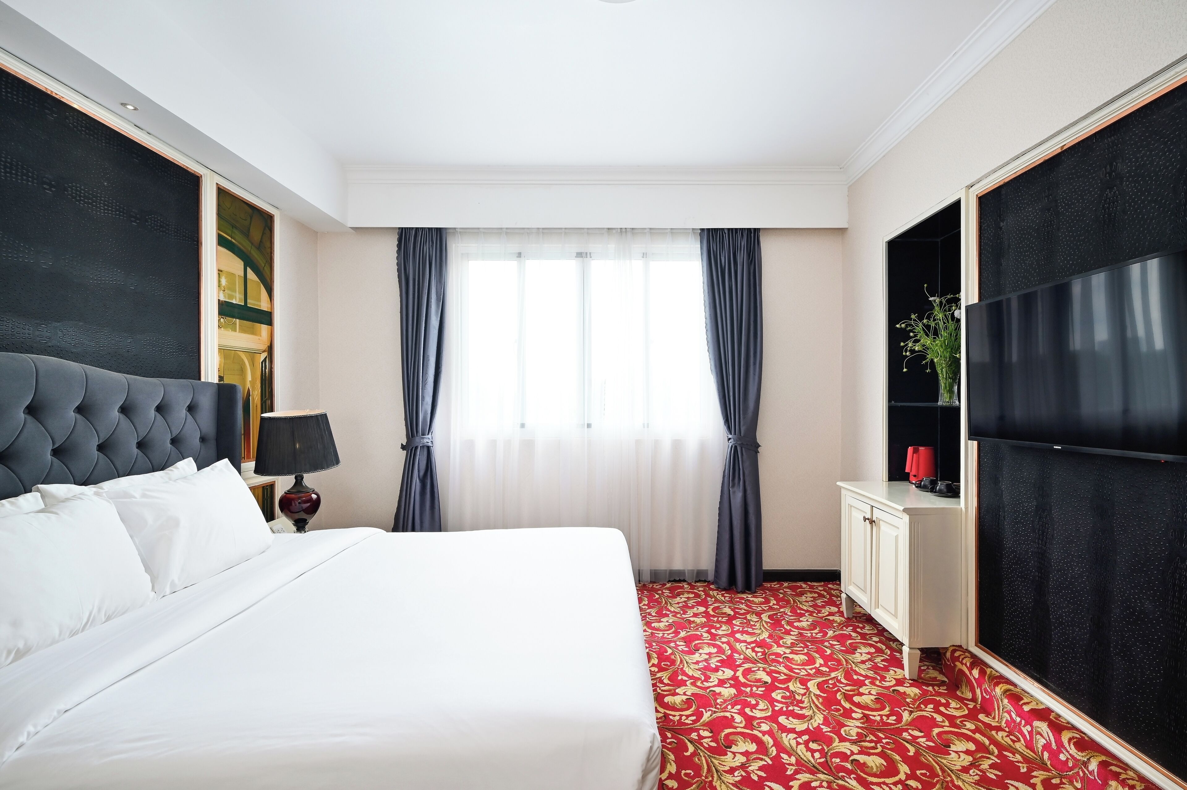 A hotel room with a large bed, a flat-screen TV, a bedside table with a lamp, and a red carpeted floor.