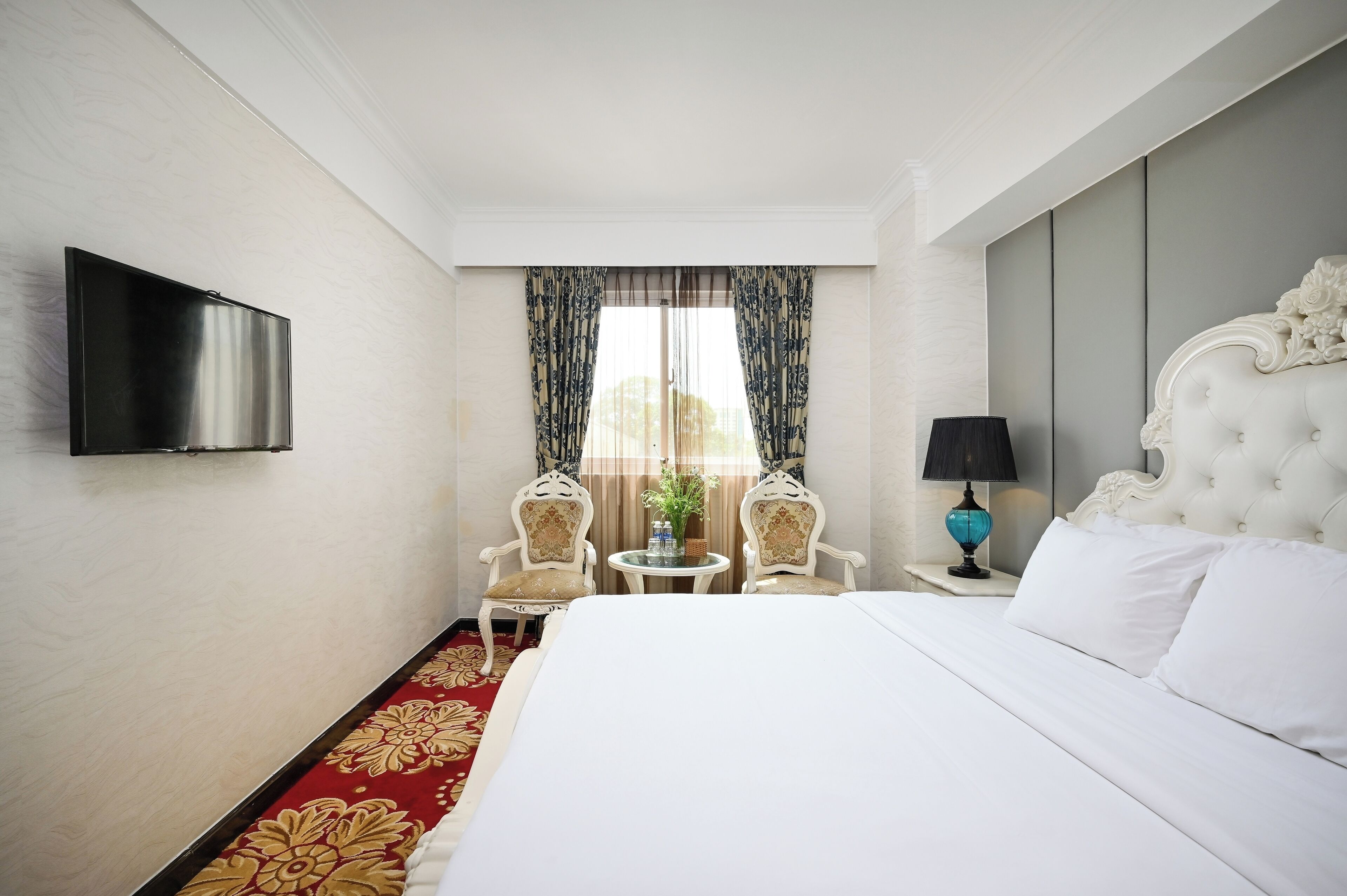 A hotel room with a large bed, a television mounted on the wall, two chairs around a small table, and a window with curtains.