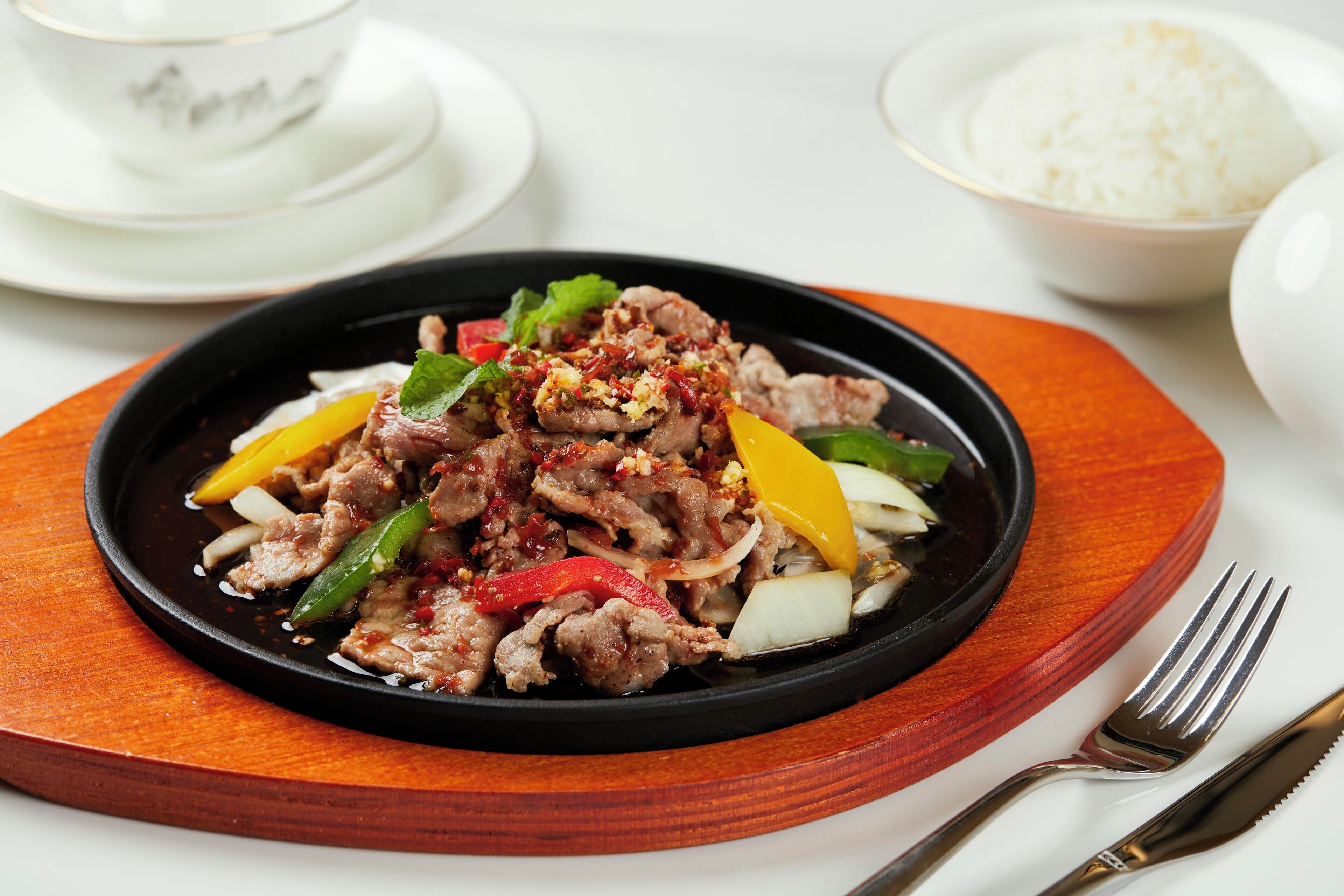 A plate of beef stir-fry with vegetables on a black plate, accompanied by a bowl of rice and a cup of tea.