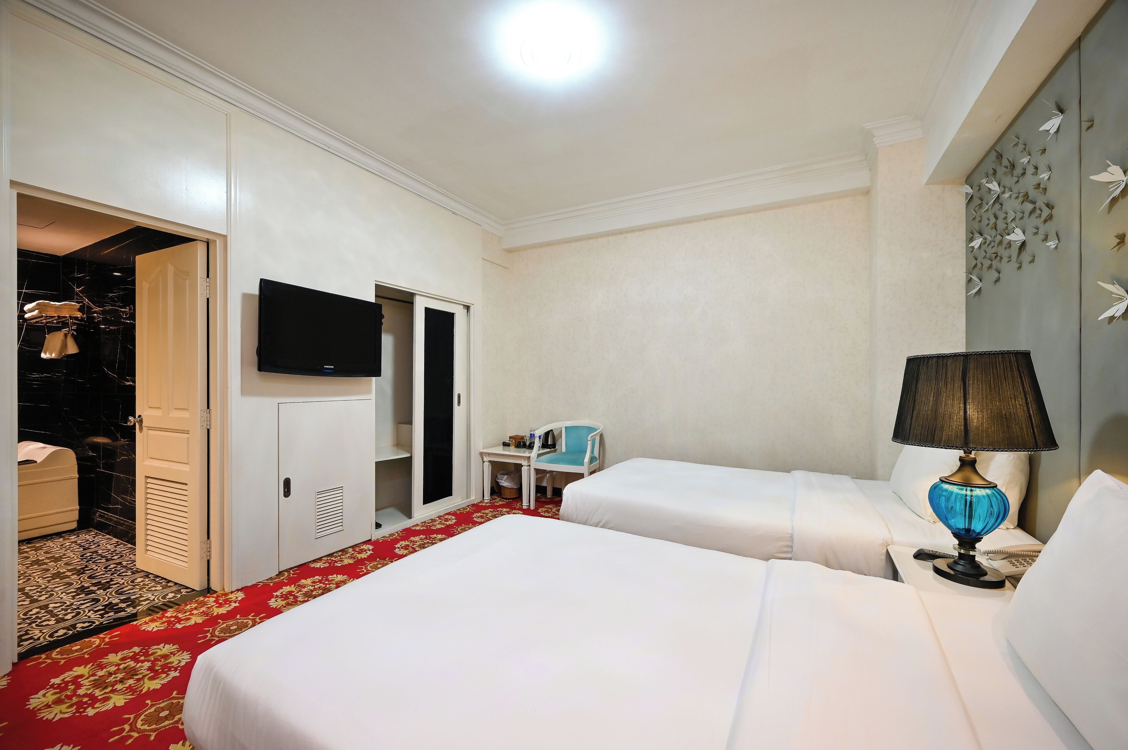 A hotel room with two beds, a TV, a desk with a chair, and a red carpet.