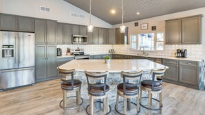 Een moderne keuken met een groot eiland, barstoelen, roestvrijstalen apparatuur en hanglampen.