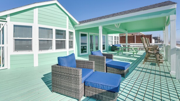 A covered outdoor seating area with wicker furniture and blue cushions, a wooden deck, and a light green building with white trim. - Great for Groups: Oceanfront Surfside Beach Home! (Freeport)