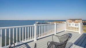 A wooden deck with white railings overlooking a body of water, a dock with boats, and a house in the background.