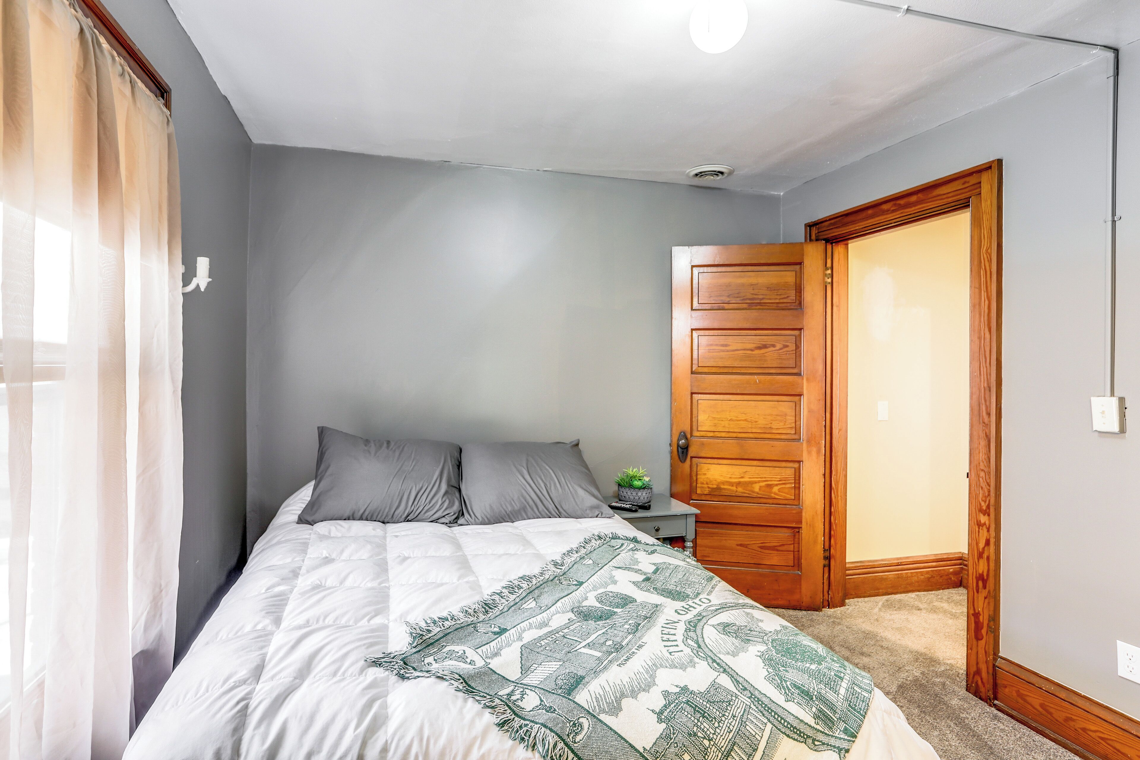A bedroom with a bed, grey walls, a wooden door, and a small nightstand with a plant.