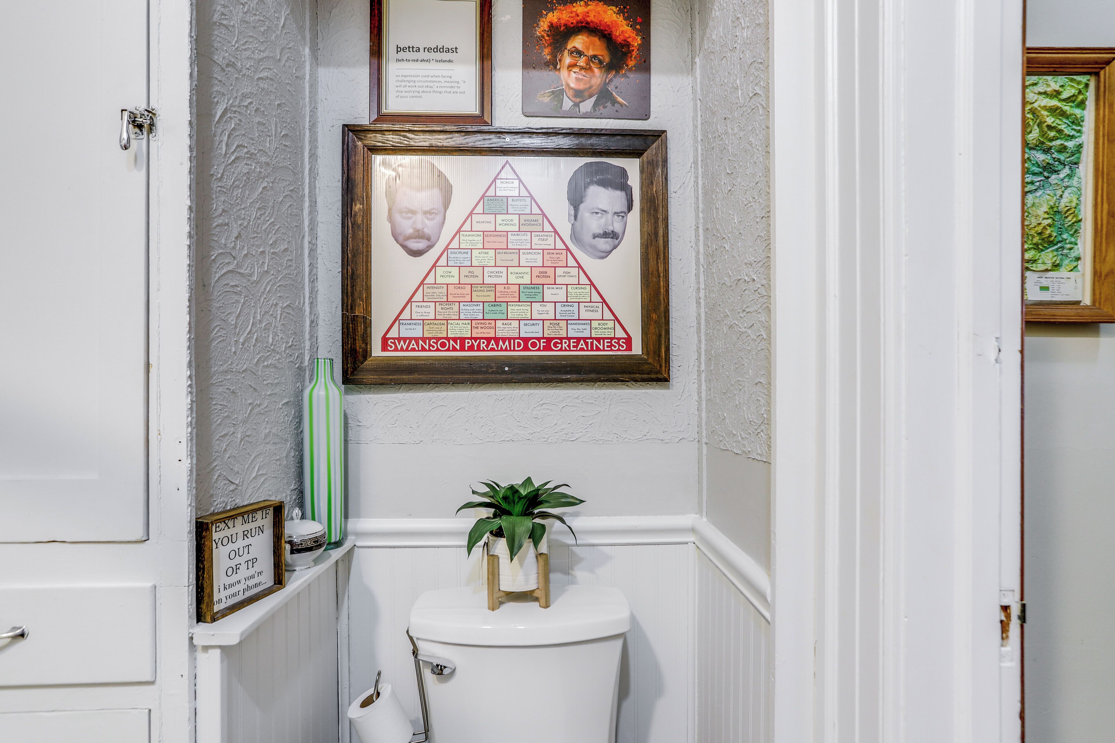 A framed Swanson Pyramid of Greatness in a bathroom with a toilet, a small plant, and a framed picture on the wall.
