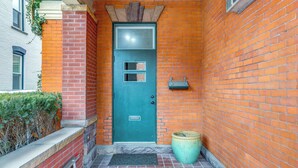 Een smalle bakstenen gang met een blauwgroene deur, een potplant en een metalen trap naar een betegelde ingang.