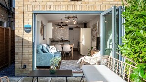 A modern outdoor living space with a sofa, dining table, and chairs, a small table with a vase, and a staircase leading to a glass door. - Stylish River Apartment - West Putney Embankment (London)