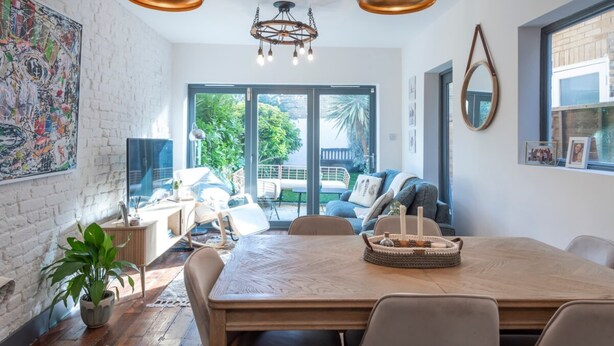 A modern dining area with a large wooden table, two hanging pendant lights, and a view of the outdoors through large sliding doors. - Stylish River Apartment - West Putney Embankment (London)