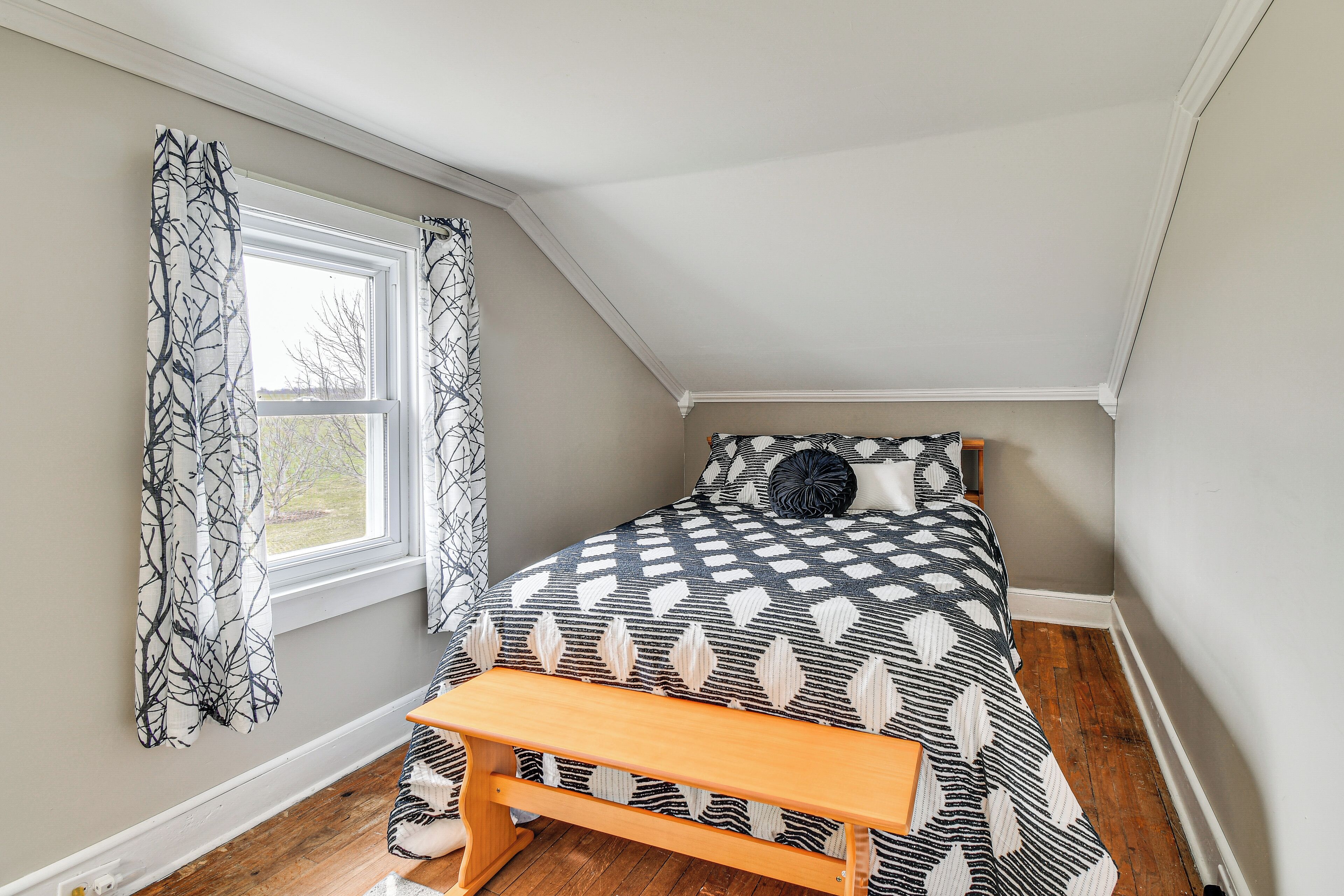 Quarto com teto inclinado, cama com colcha estampada, um banquinho e cortinas.