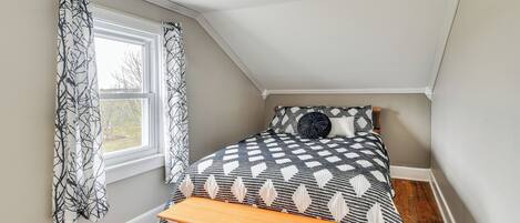 Quarto com teto inclinado, cama com colcha estampada, um banquinho e cortinas.