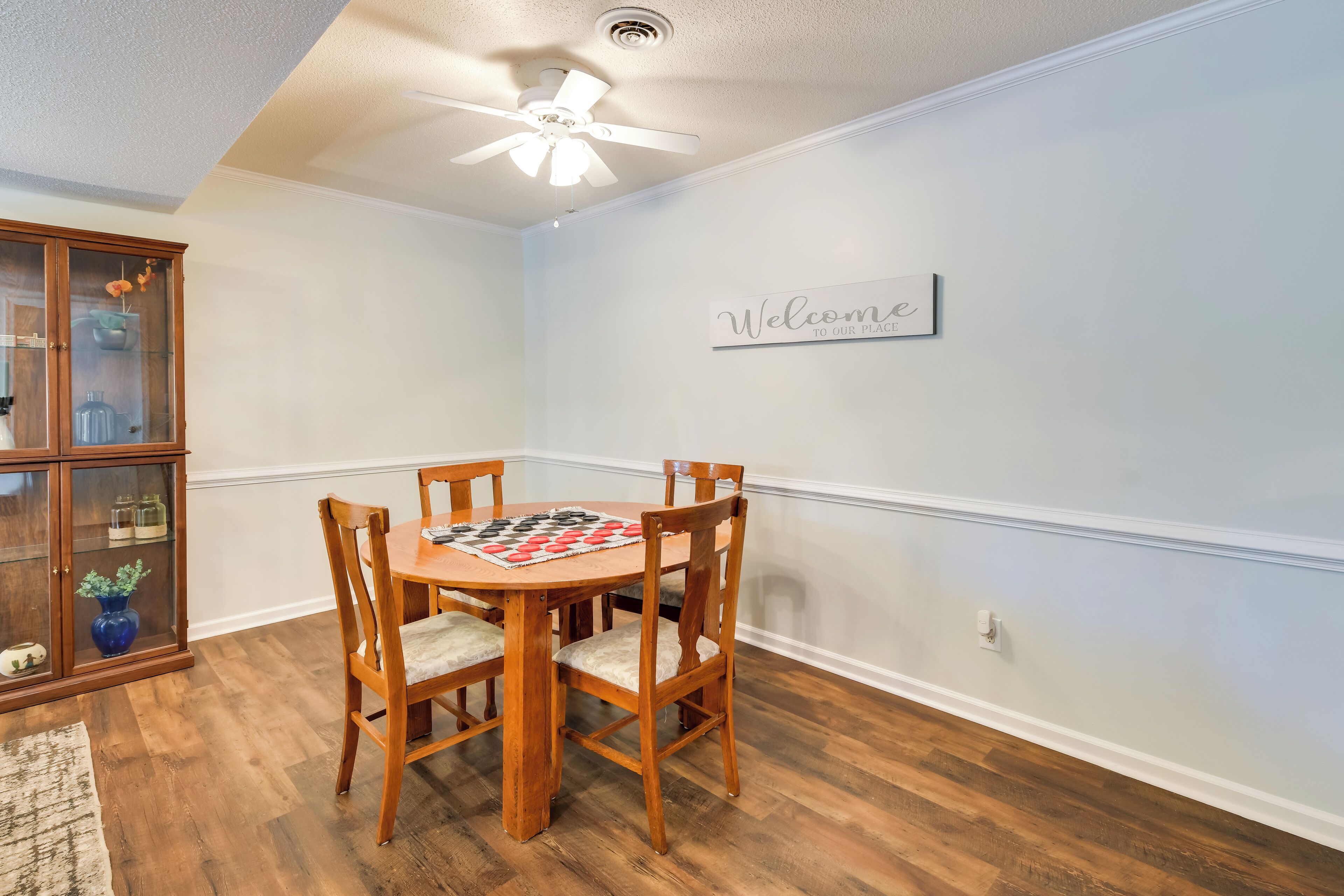 A dining area with a round wooden table, four chairs, a chessboard, a cabinet with glass doors, and a 'Welcome' sign on the wall.
