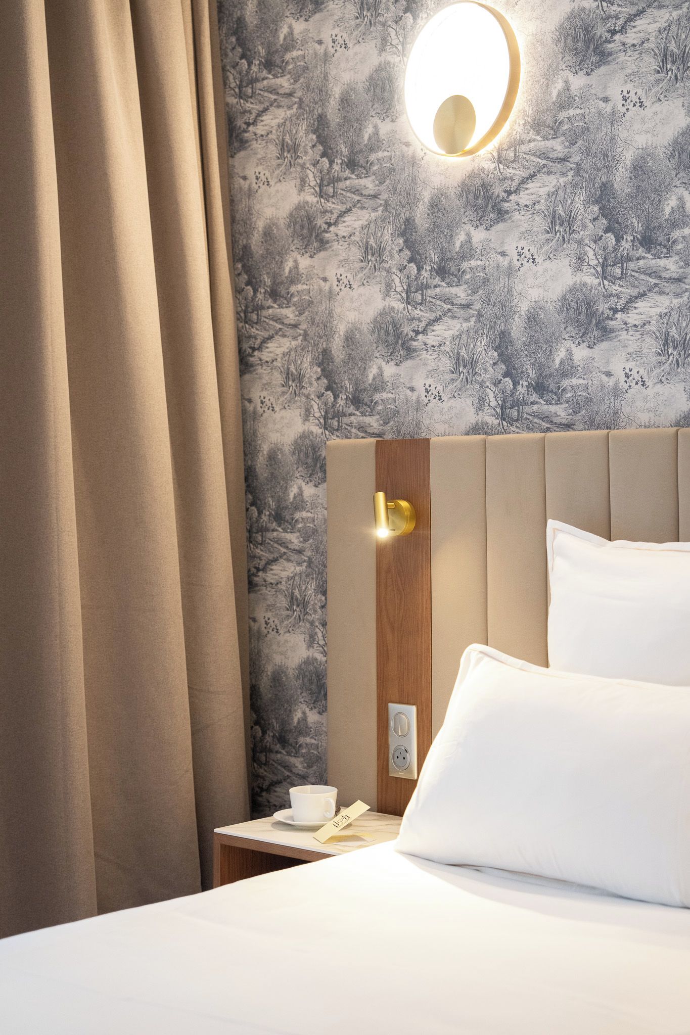 A hotel room with a bed, a nightstand, a cup, and a wall-mounted light fixture.