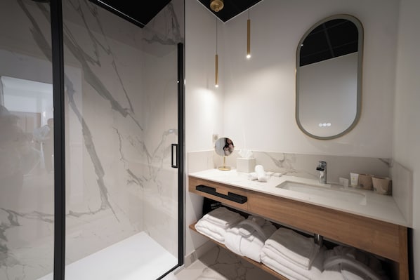 Un baño moderno con cabina de ducha de vidrio, pared con azulejos de mármol, tocador de madera con lavamanos y espejo con marco dorado.