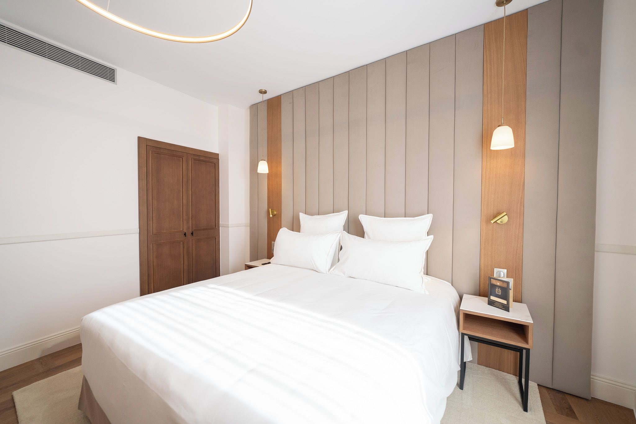 A hotel room with a large bed, a wooden wardrobe, a bedside table with a lamp, and a wall-mounted light fixture.