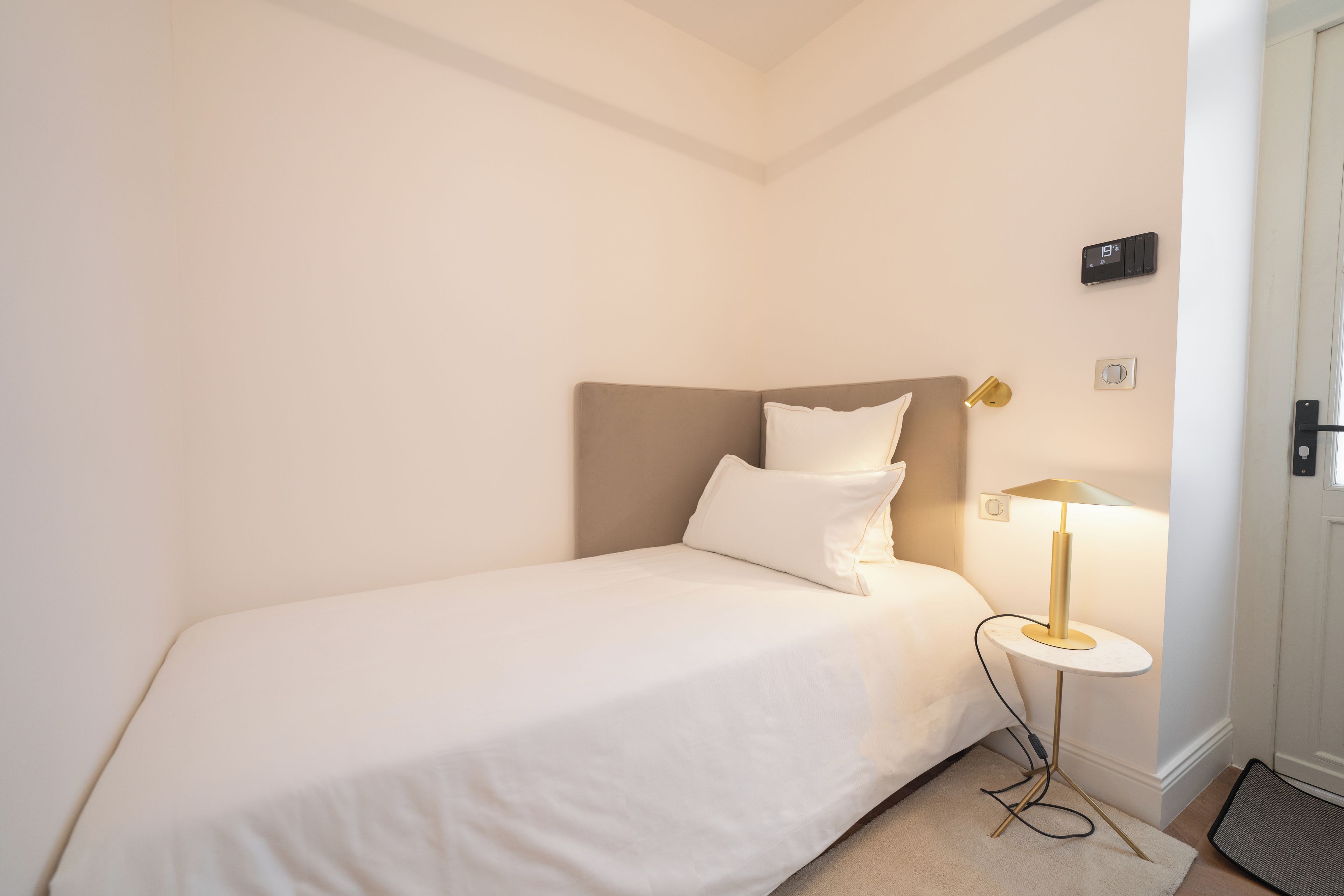 A single bed with a headboard, a bedside lamp, a wall clock, and a door with a handle.