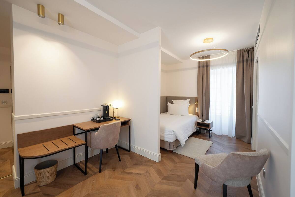 A hotel room with a bed, a desk with a chair, a small table, and a lamp.