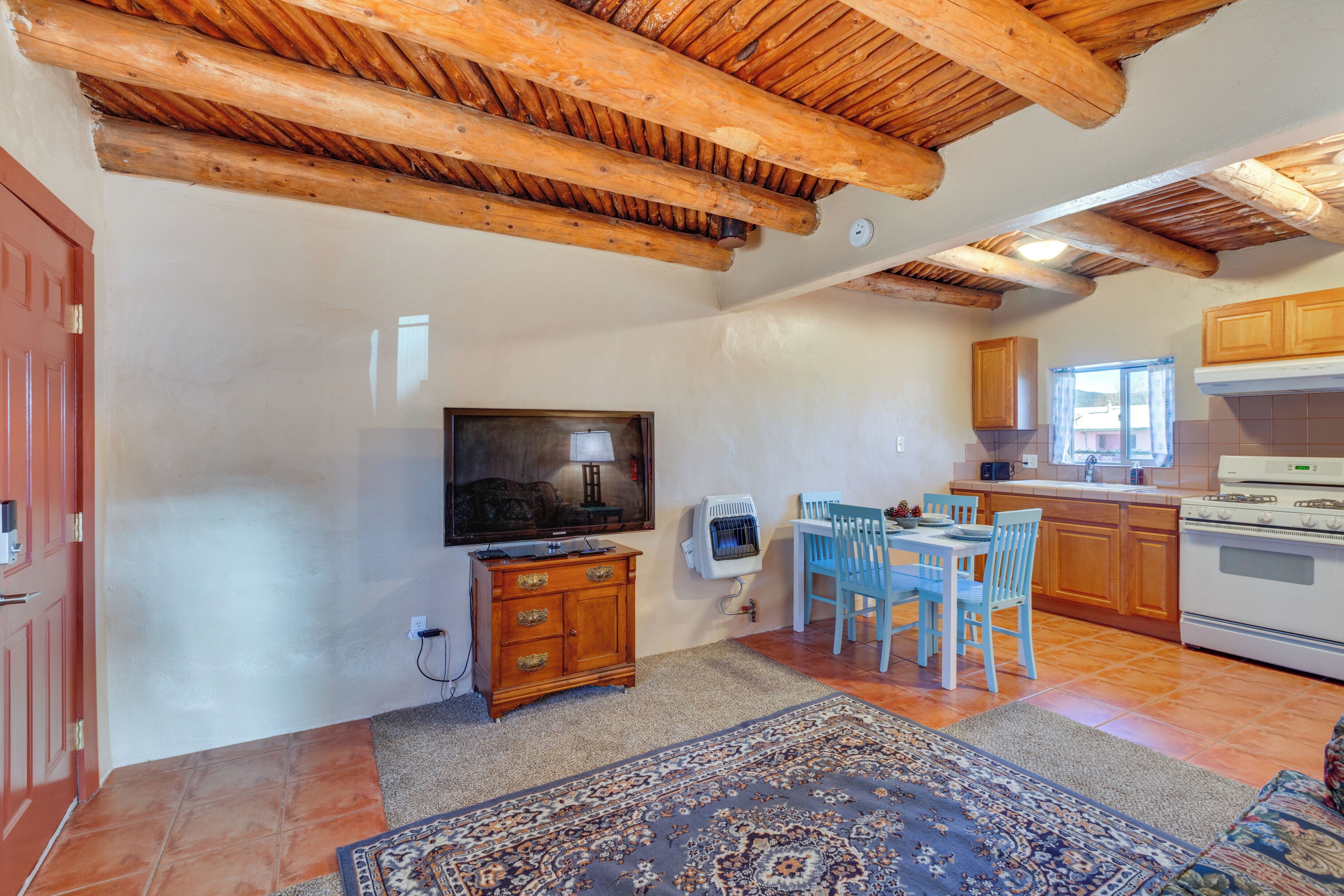 A kitchen with wooden beams, a dining area with blue chairs, a TV on a wooden cabinet, and a tiled floor. - Quiet Mora Hideaway: 35 Mi to Angel Fire Resort! (Mora)