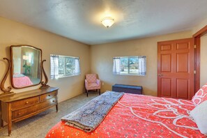 Quarto com cômoda de madeira, cama com colcha florida vermelha, um banquinho e duas janelas com cortinas.