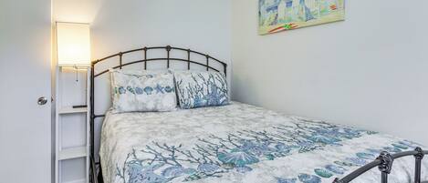 A bed with a metal frame, a white door, a wall-mounted shelf, and a painting of a beach scene.