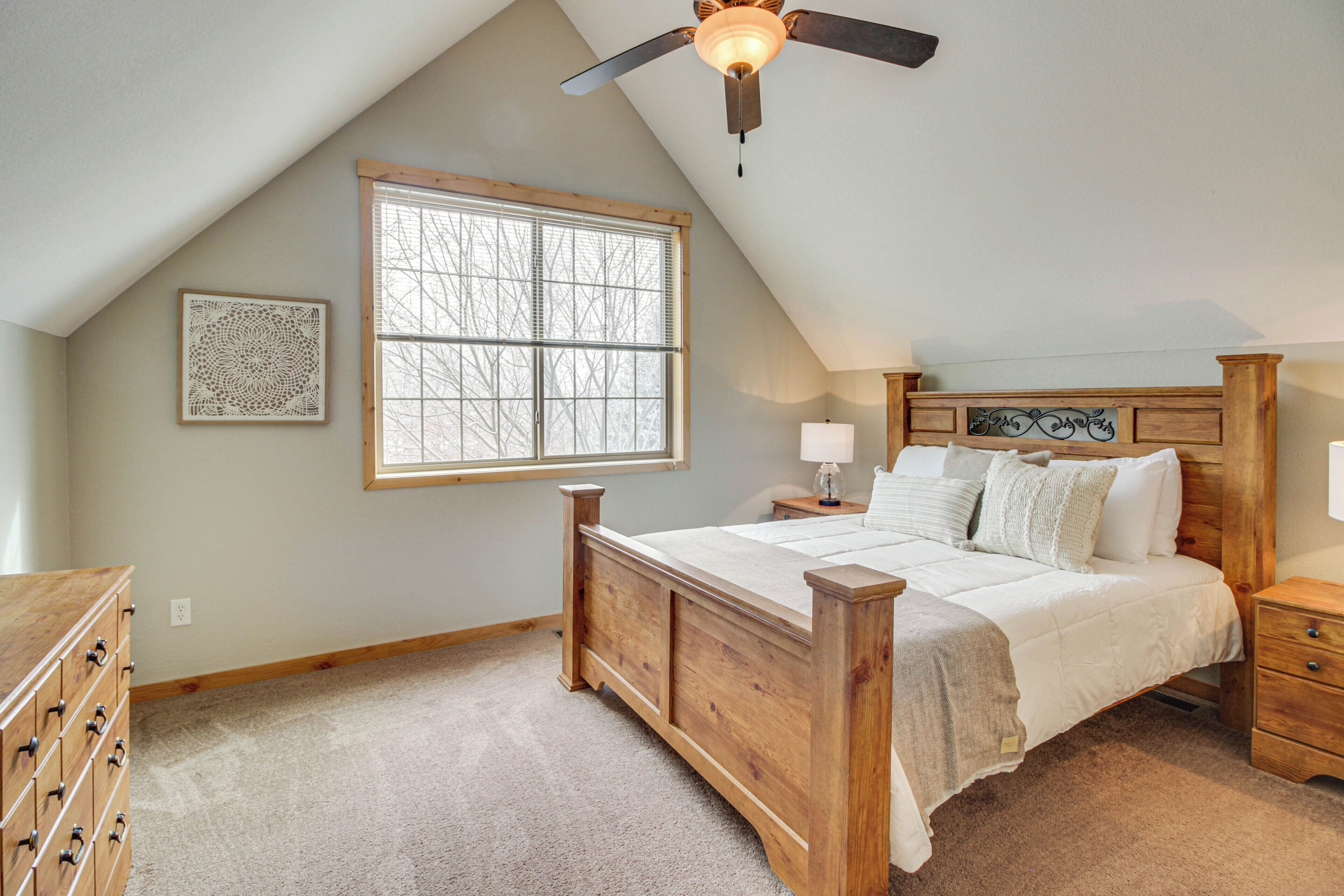 A bedroom with a sloped ceiling, a large window, a wooden bed, a nightstand, a dresser, and a ceiling fan.