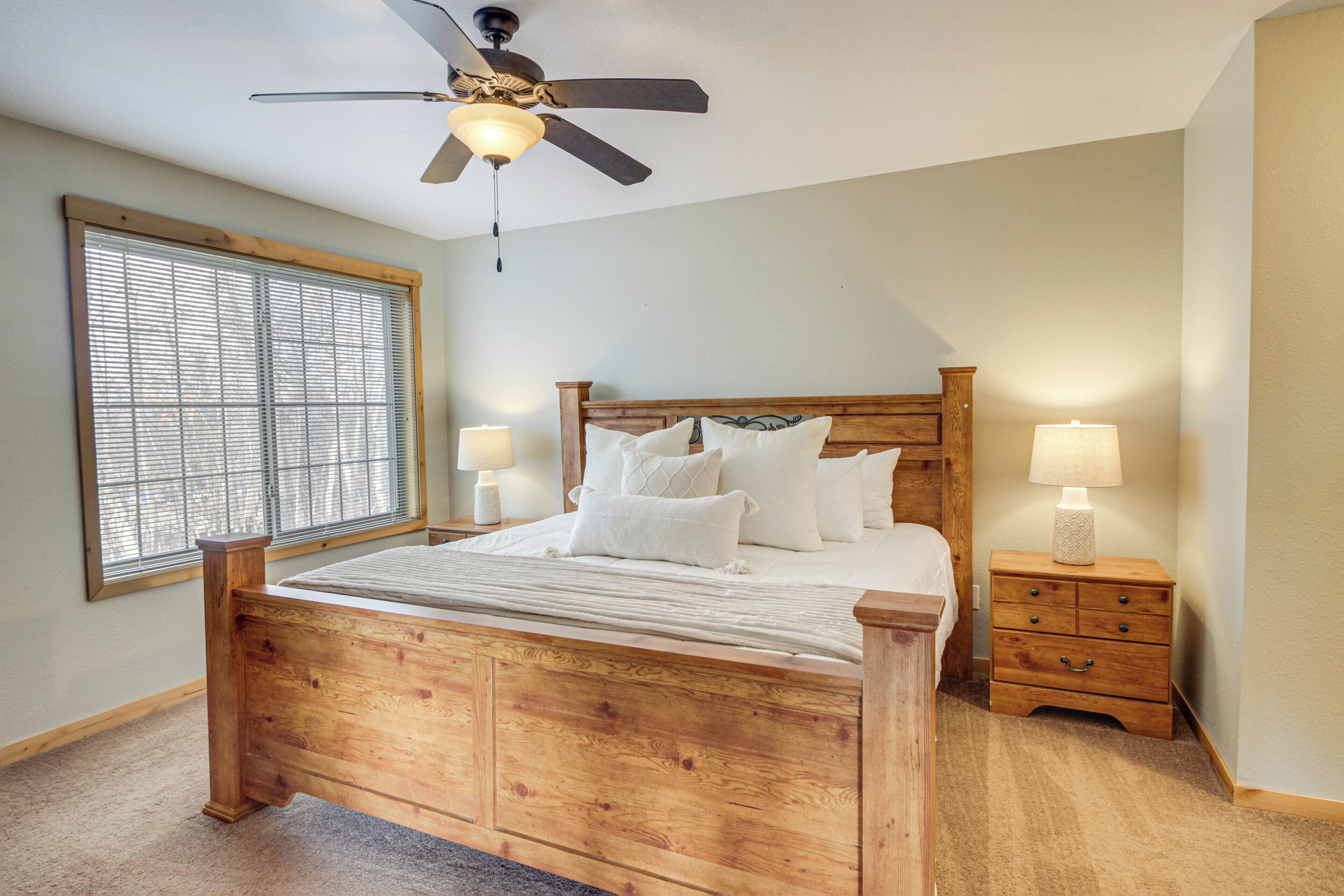 Un dormitorio con cama de madera, ventilador de techo, dos mesitas de noche con lámparas y una ventana con persianas.